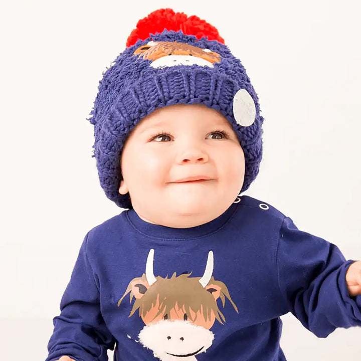 Child wearing a Blade & Rose Hamish Highland Cow blue knit hat with a red pom-pom and a matching blue shirt with a cow design on a white background