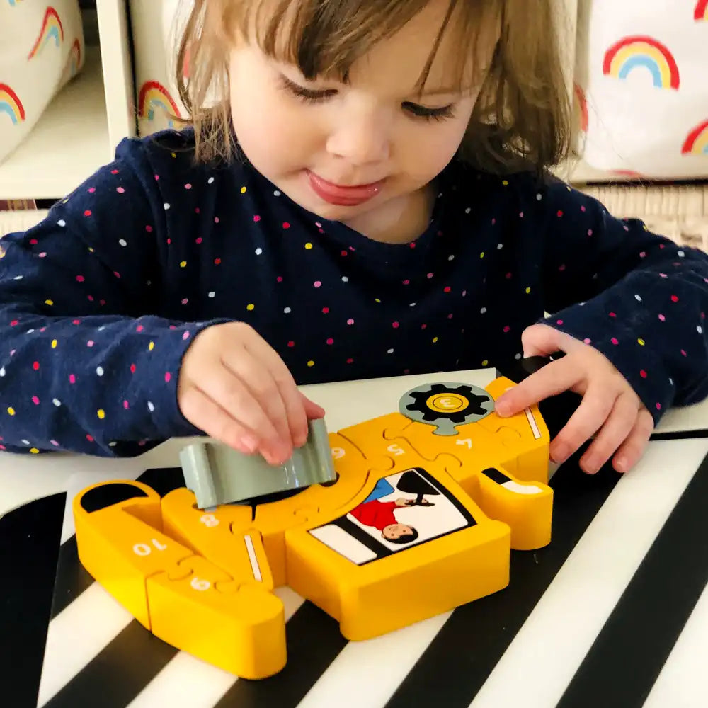 A young child wearing a navy blue long-sleeve shirt with colourful polka dots is playing with a Lanka Kade yellow toy digger on a black and white striped table.