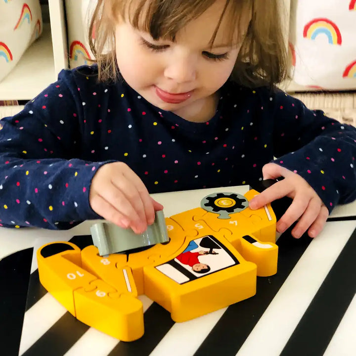 A young child wearing a navy blue long-sleeve shirt with colourful polka dots is playing with a Lanka Kade yellow toy digger on a black and white striped table.