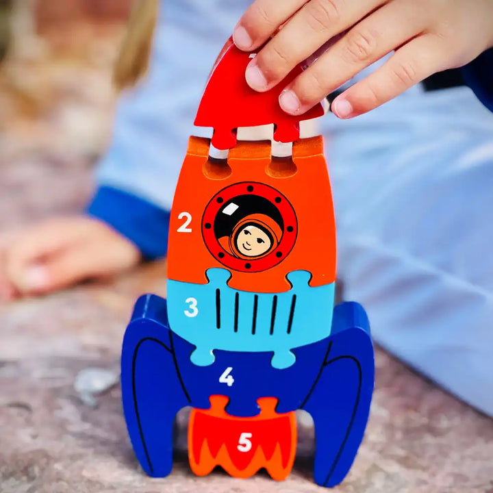 Child's hand holding a colourful Lanka Kade rocket-shaped puzzle with numbers on a wooden surface.
