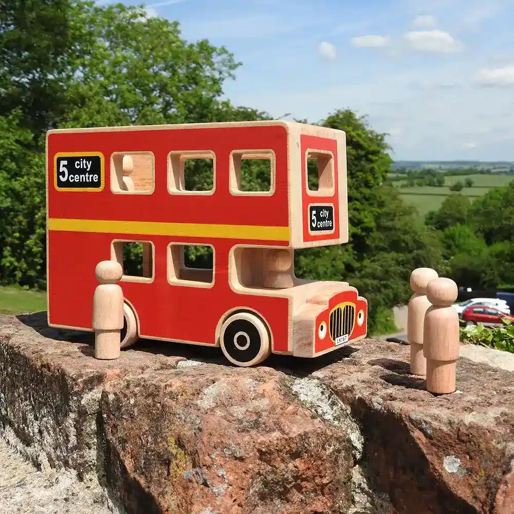 A Lanka Kade wooden double-decker toy bus in red with natural wood trim and black wheels, displayed outdoors on a stone ledge with small wooden peg people around it. Trees and a blue sky are visible in the background.