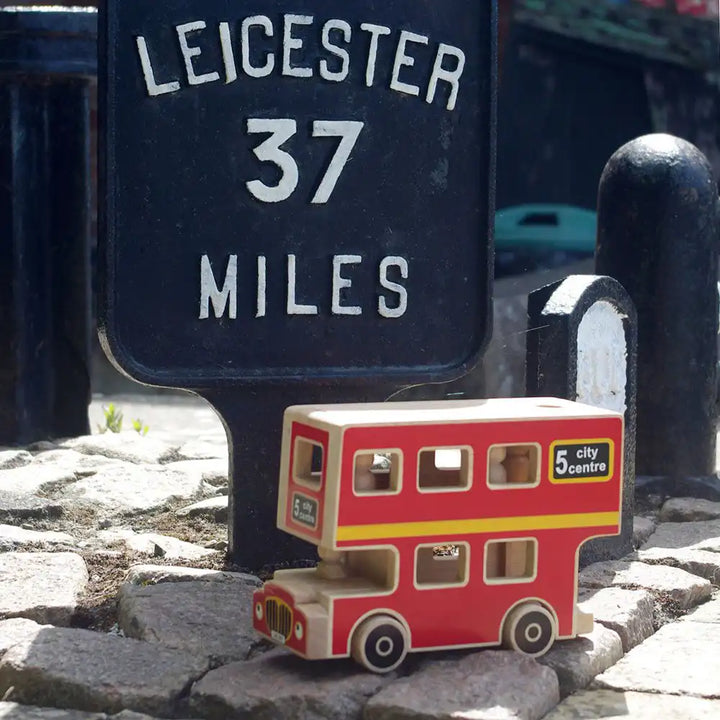 A Lanka Kade small wooden toy double-decker bus in red, cream, and yellow sits on cobblestones in front of a black Leicester 37 Miles sign.