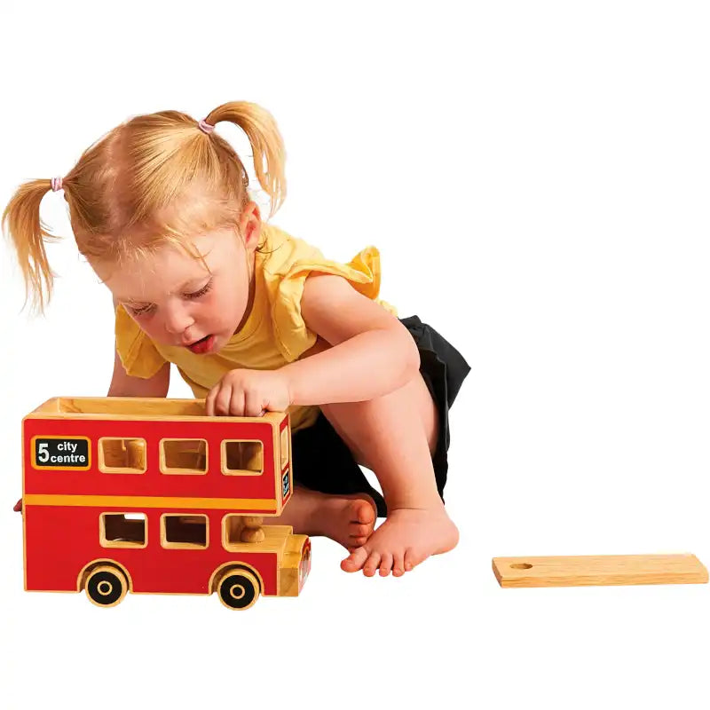A toddler girl with blonde pigtails in a yellow top and black skirt plays with a red and yellow Lanka Kade wooden city bus playset on a white background. A small wooden piece lies nearby.