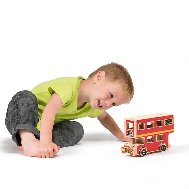 A young boy in a lime green t-shirt and dark grey pants sits on the floor looking at a Lanka Kade red wooden city bus playset with cut-out windows.