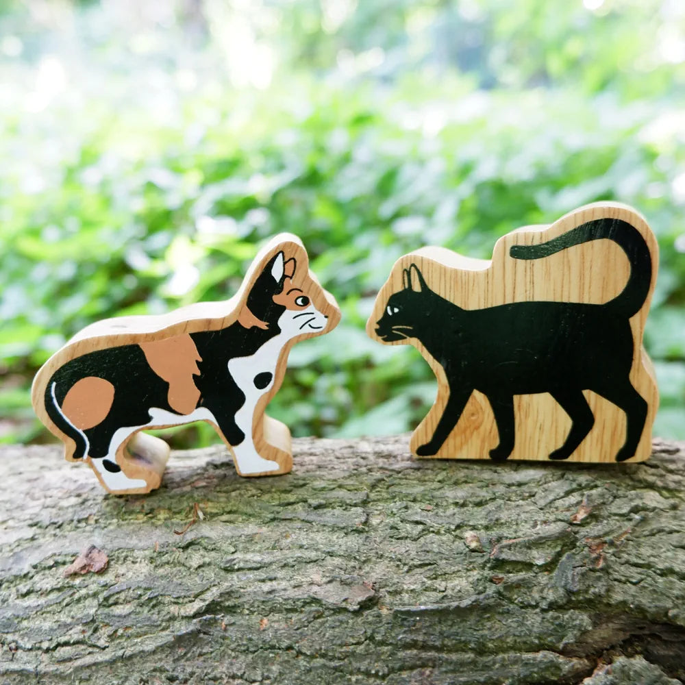 Two Lanka Kade wooden animal toy figures, one tabby cat and one black cat, displayed on a tree trunk with a blurred green forest background.