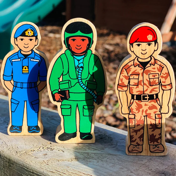 Three Lanka Kade wooden military-themed toy soldiers standing on a weathered wooden surface outdoors. The left soldier is in a blue navy uniform, the middle soldier is in a green air force suit, and the right soldier is in a brown camo military uniform.