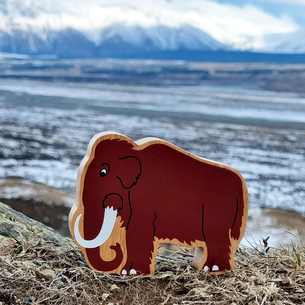Lanka Kade wooden toy mammoth, featuring a reddish-brown painted body with natural light wood edges, black line details, standing on a log in front of a blurred outdoor landscape with mountains and snow.
