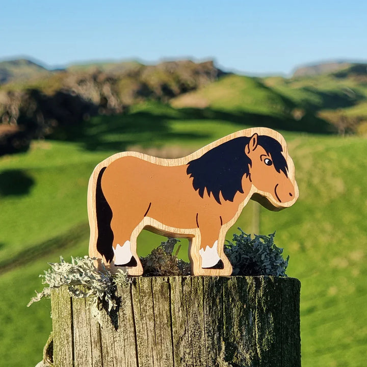 Lanka Kade wooden Shetland pony toy with brown body, black mane, and white feet, displayed on a weathered wooden post with moss in a blurred green outdoor landscape.