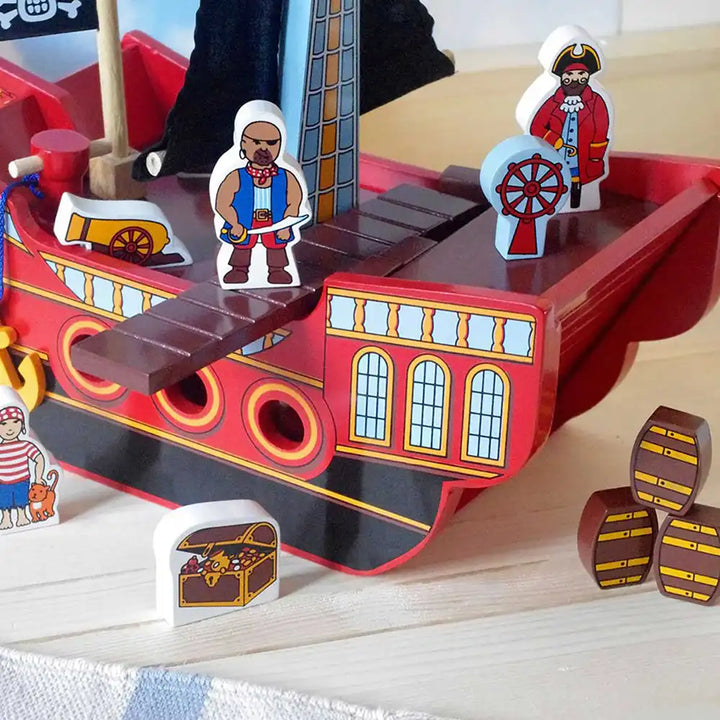 Lanka Kade red and black pirate ship toy with colourful wooden pirate figures, a treasure chest, barrels, and a wheel on a light wood tabletop.