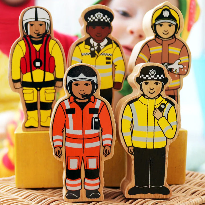 A set of Lanka Kade wooden toy figures representing firefighters, police officers, and an emergency worker, standing on yellow blocks against a softly blurred background.