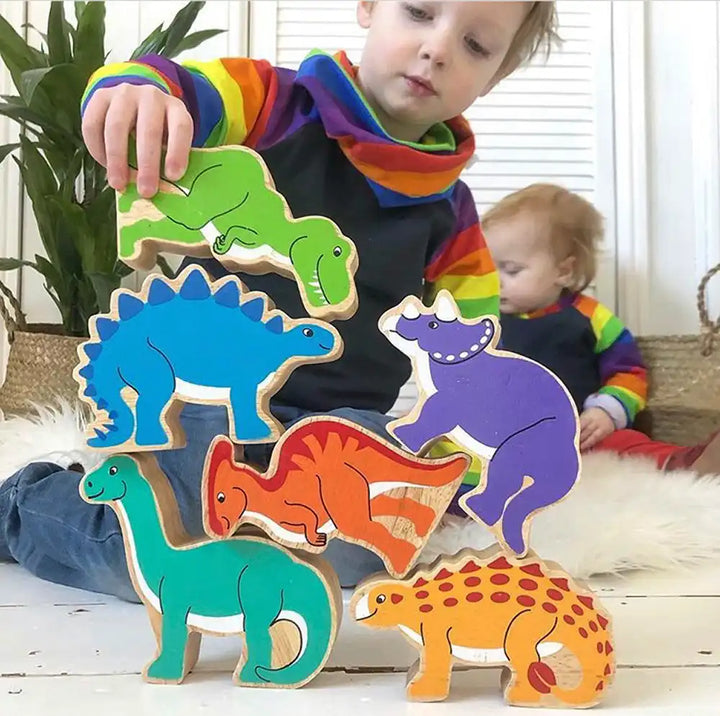 A child's Lanka Kade wooden dinosaur puzzle toy set with colourful dinosaur cut-outs, shown being assembled by a young child on a white floor with a plant in the background.