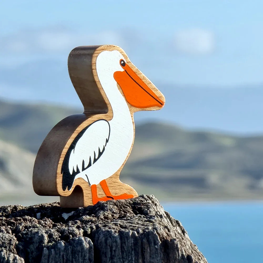 Lanka Kade wooden pelican, featuring a natural light wood body, a white head and body, orange beak and legs, and black wing details, standing on a dark rock with a blurred blue water and mountain background.