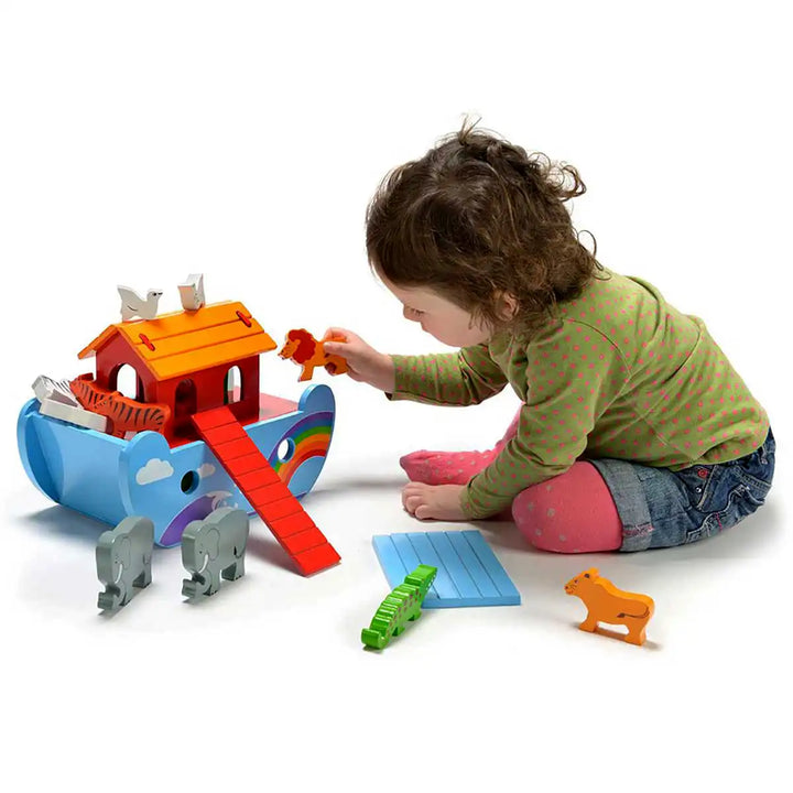 A toddler sits on the floor playing with a colourful wooden Lanka Kade Noah's Ark toy set, including a blue ark, orange roof, red ramp, grey elephants, green caterpillar, and orange animal pieces on a white background.