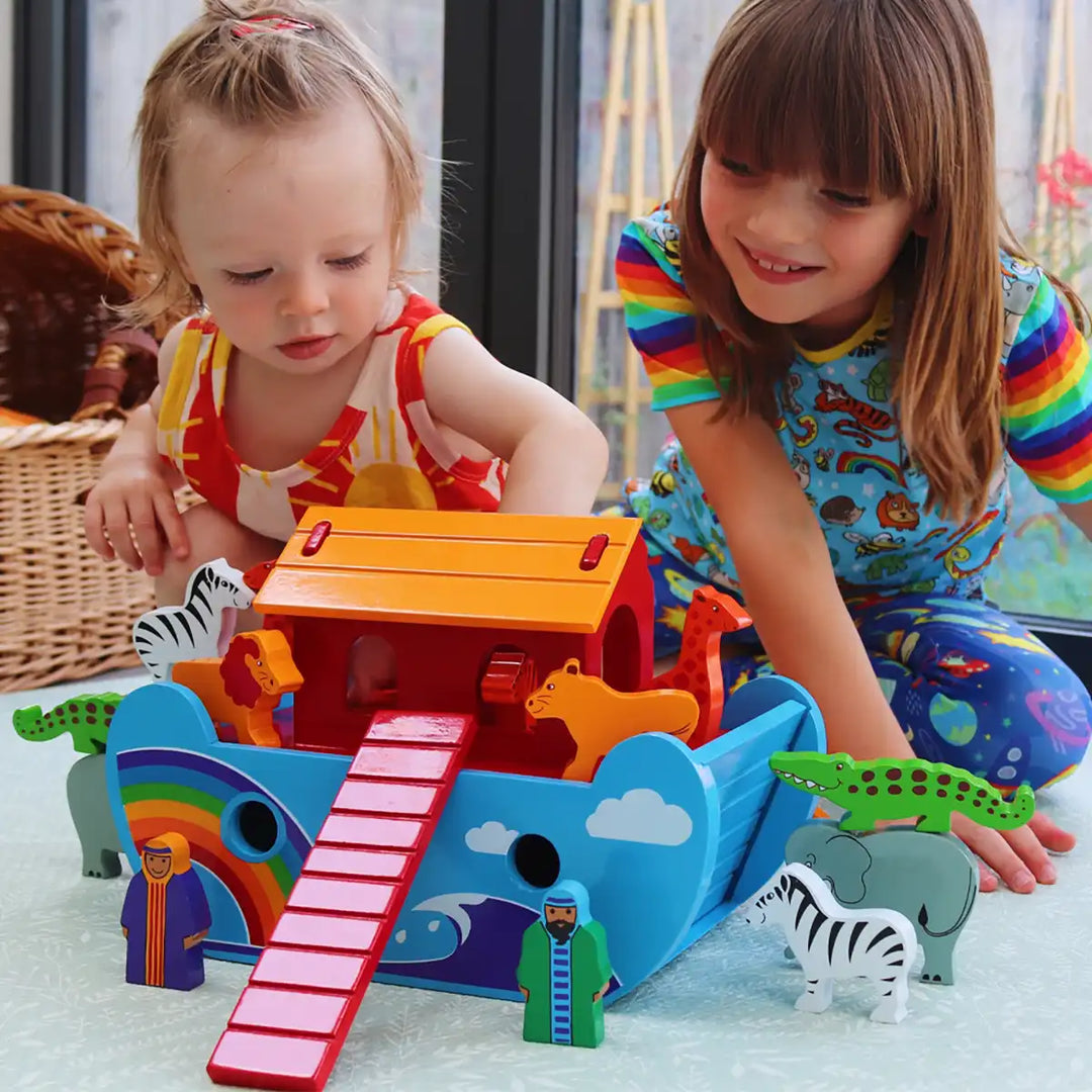 Two young children playing with a colourful wooden Lanka Kade Noah's Ark toy set on a light surface indoors. The toy includes a blue ark with a red ramp, orange roof, rainbow panel, and small animal figures and blocks.