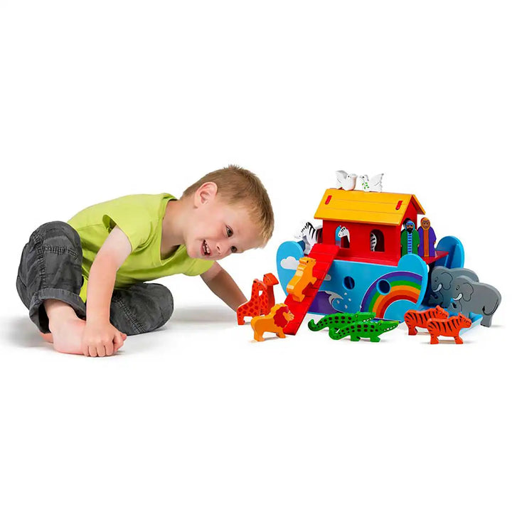 A young child in a lime green shirt and dark pants lies on a white floor, smiling at a colourful wooden Lanka Kade toy Noah's Ark playset with animal figures and a rainbow.