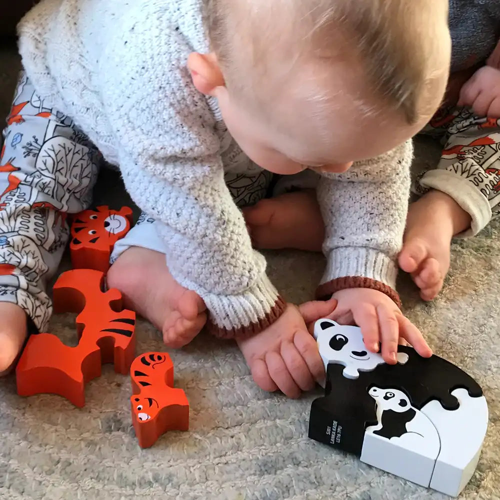 A baby in a white knit sweater crawls on a carpeted floor, touching a Lanka Kade black-and-white panda puzzle piece. Several orange wooden animal puzzle pieces are scattered nearby, including tiger and lion shapes.
