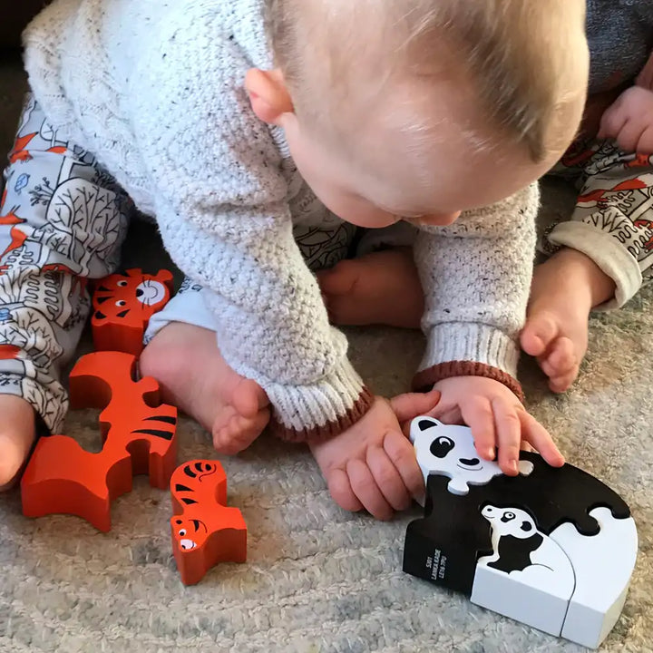 A baby in a white knit sweater crawls on a carpeted floor, touching a Lanka Kade black-and-white panda puzzle piece. Several orange wooden animal puzzle pieces are scattered nearby, including tiger and lion shapes.