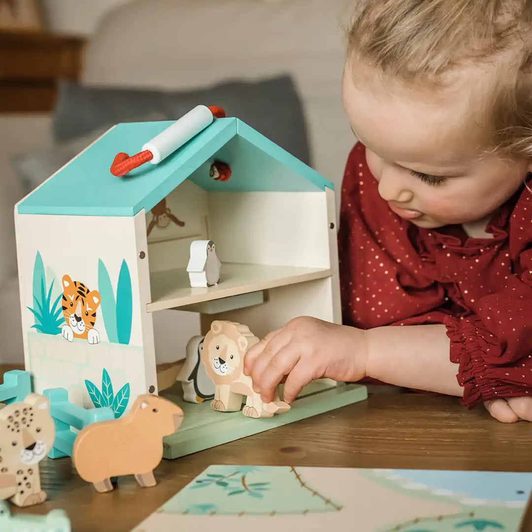 Child playing with an Orange Tree Toys wooden zoo play set with house and animal figures on a table.