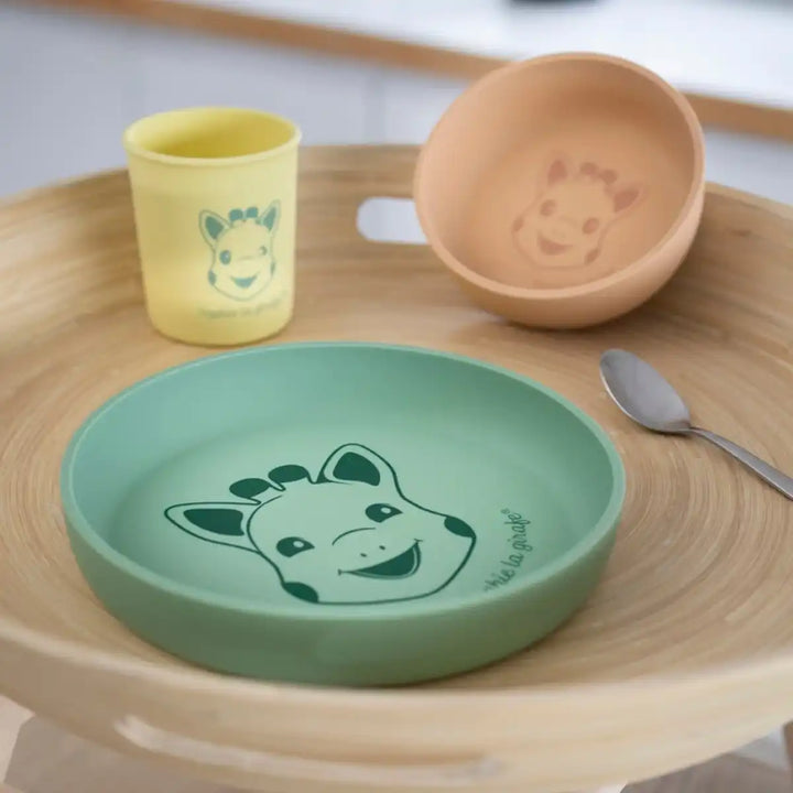 Sophie La Girafe silicone children's tableware set with green plate, yellow cup, and brown bowl on a wooden surface.