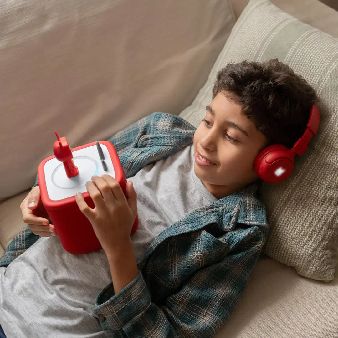 Child wearing Tonies red headphones and holding a sunset red Toniebox 2  console on a couch.